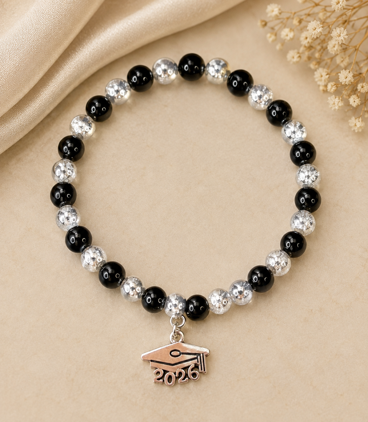 A beaded bracelet with alternating black and silver glittery beads, featuring a small graduation cap charm engraved with '2026', displayed on a beige fabric background with dried flowers.