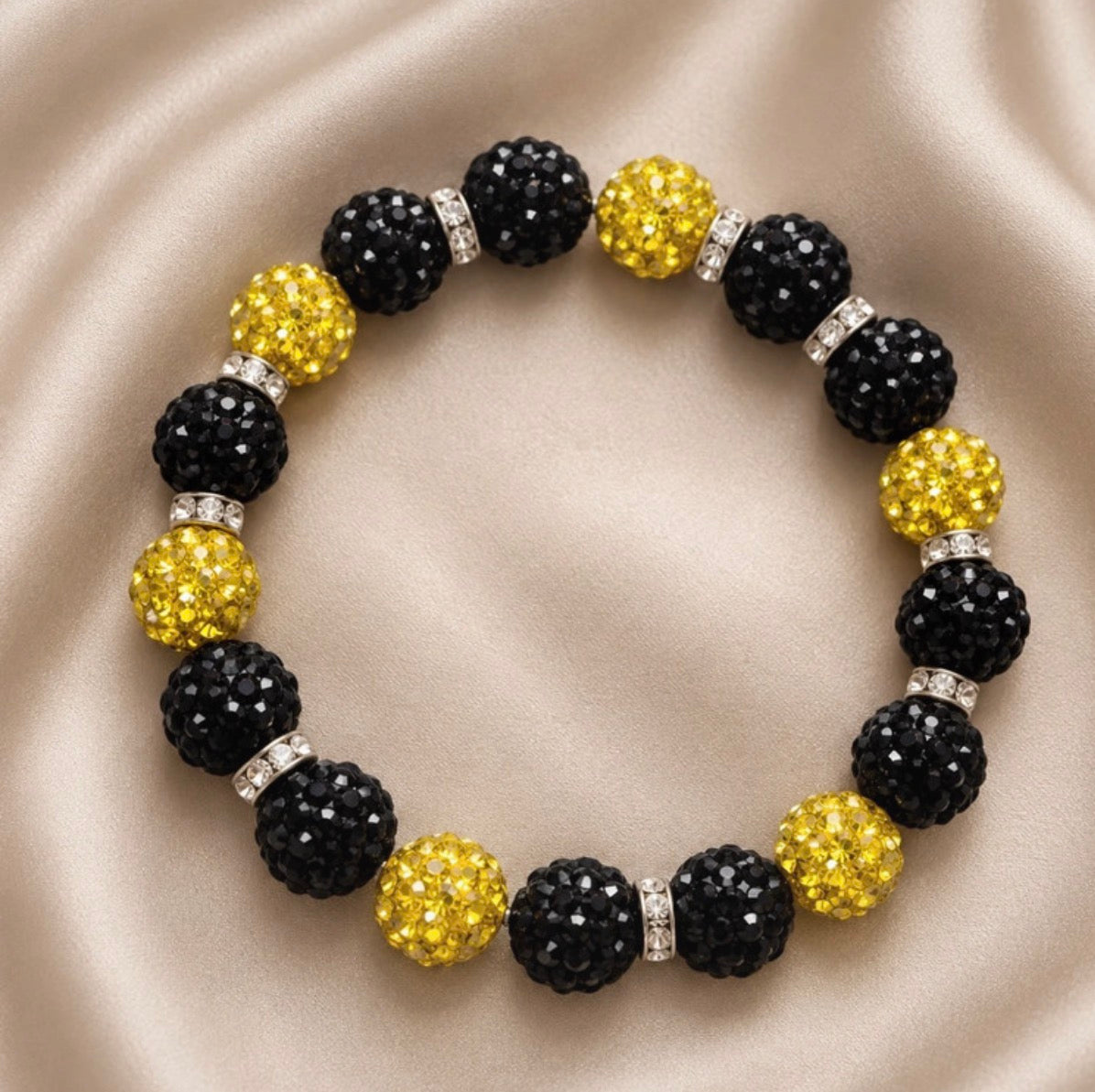 A beaded stretch bracelet featuring alternating black faceted crystal beads and yellow faceted crystal beads, separated by small iridescent spacers, displayed on a beige satin fabric background.
