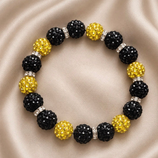 A beaded stretch bracelet featuring alternating black faceted crystal beads and yellow faceted crystal beads, separated by small iridescent spacers, displayed on a beige satin fabric background.