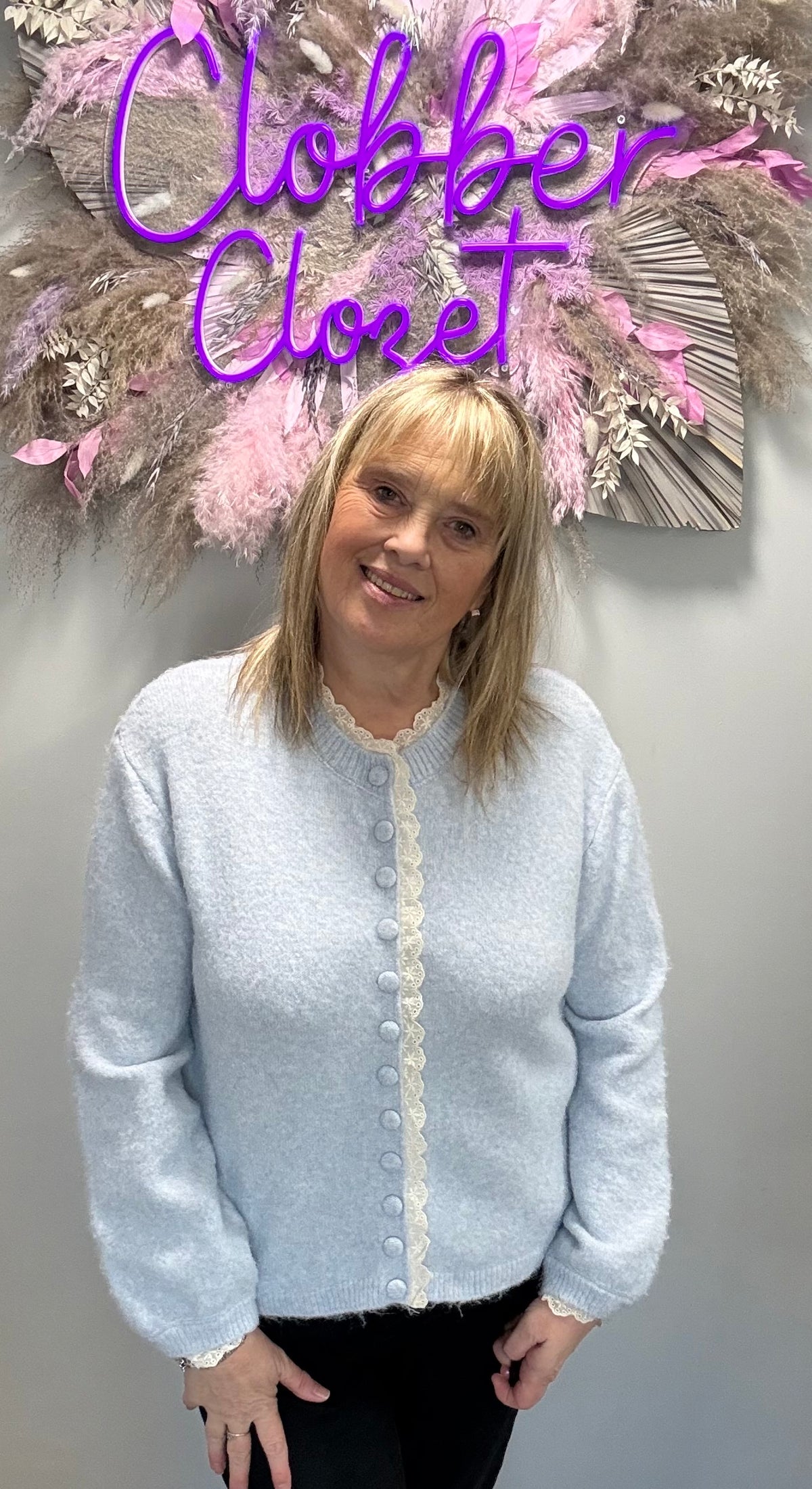 A woman wearing a light blue fuzzy knit cardigan with a cream lace collar and button-front detail, standing in front of a gray wall with a decorative floral wall hanging and purple 'Closet' text.
