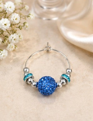 A silver wine glass charm with a central round blue glitter bead and two smaller blue and silver beads, displayed on a beige surface with small white flowers and a soft-focus glass in the background.