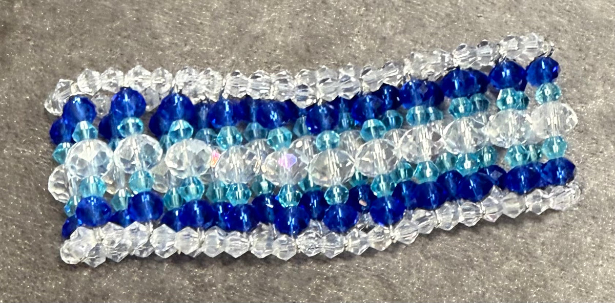 A handmade beaded bracelet featuring rows of faceted beads in royal blue, light blue, and clear/white, laid flat on a textured gray surface.