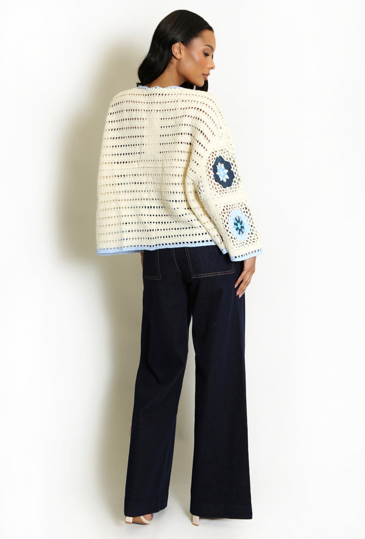 Back View of Woman wearing a Blue and Cream Open Floral Crochet Cardigan with blue patterns and black pants on a white background