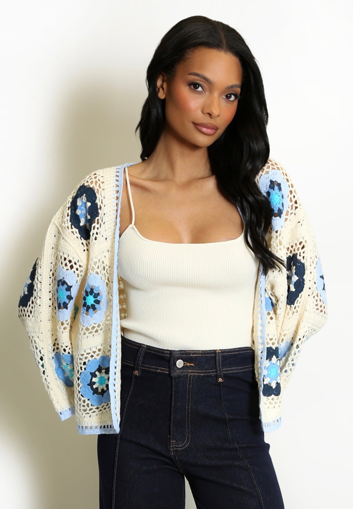Woman wearing a Blue and Cream Open Floral Crochet Cardigan over a white tank top and dark jeans on a plain background