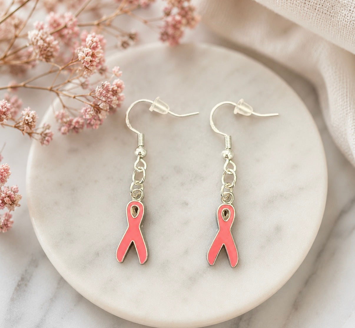 A pair of silver-tone drop earrings with pink ribbon charms, displayed on a round white marble coaster with dried pink flowers and a beige fabric background.