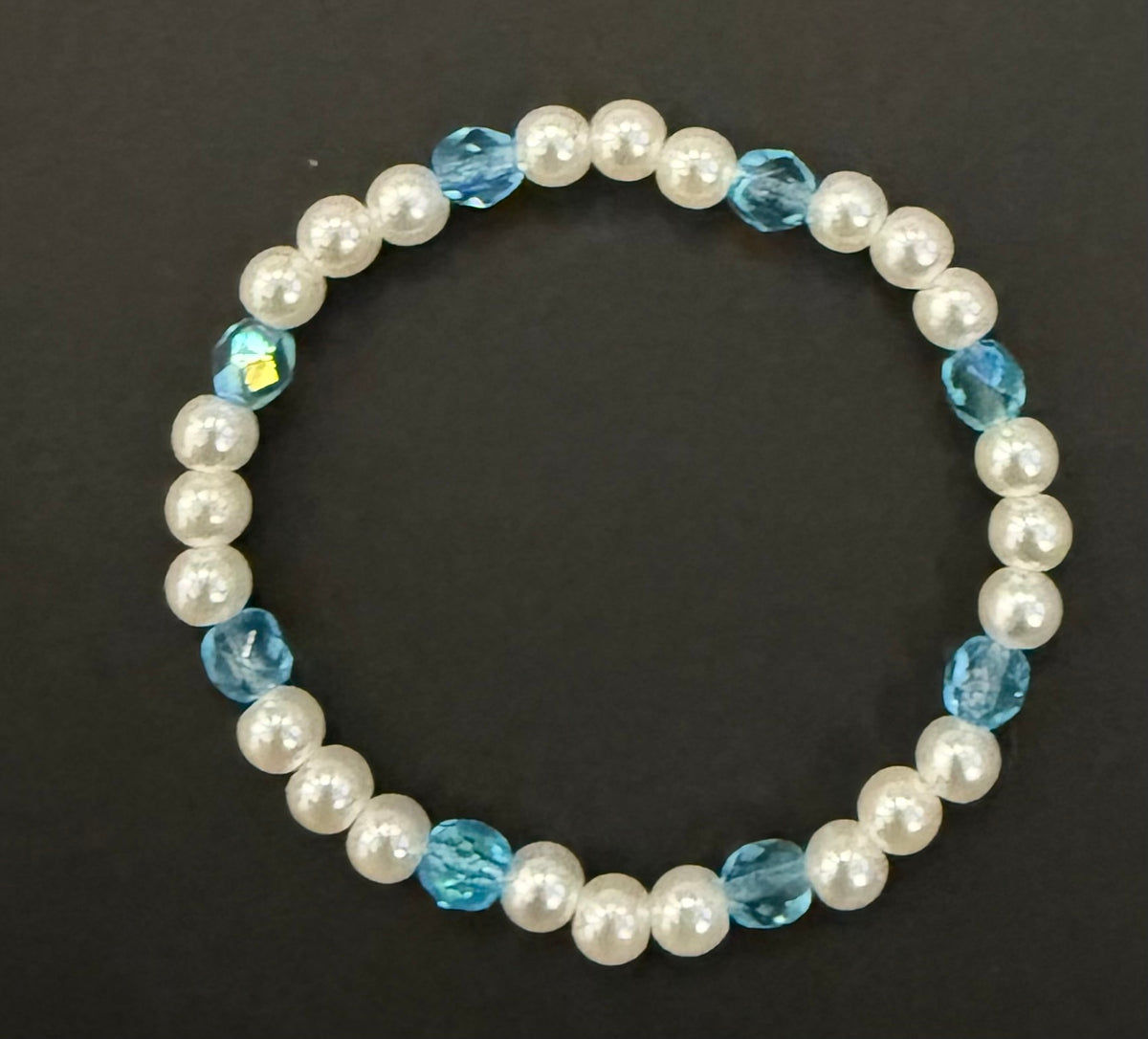 A beaded stretch bracelet featuring white faux pearls and faceted light blue beads, displayed on a dark background.