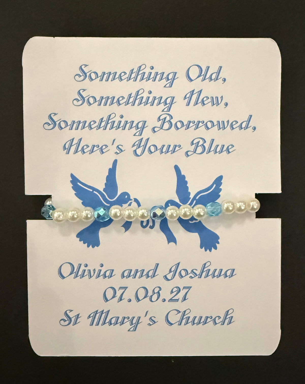 A beaded bracelet with white faux pearls and light blue faceted beads, displayed on a white printed card with blue text and a blue dove design. The card reads, 'Something Old, Something New, Something Borrowed, Here's Your Blue' and includes the names, date and venue.