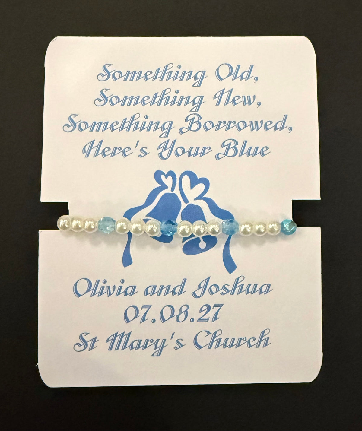 A personalized beaded bracelet with white pearls and light blue beads, displayed on a white printed card with blue text and a blue bell graphic. The card reads 'Something Old, Something New, Something Borrowed, Here's Your Blue' and names, date and venue. 