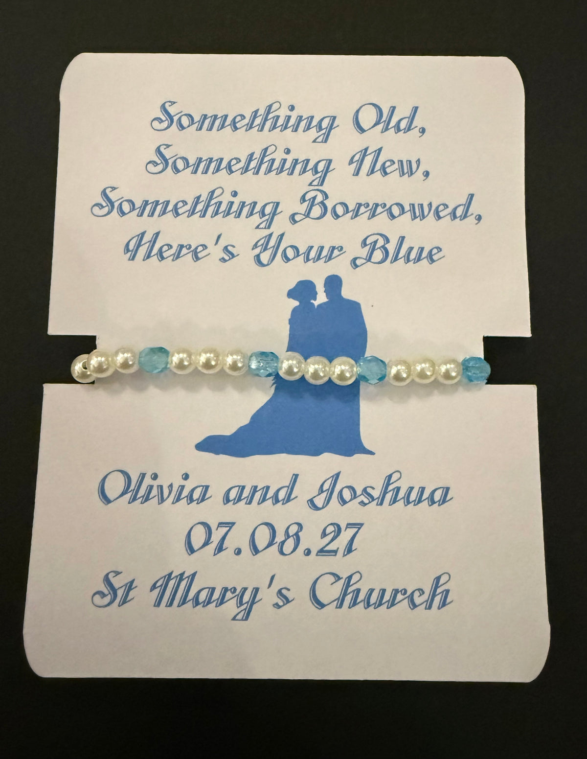 A personalized beaded bracelet with white pearls and light blue beads, displayed on a white card with blue text and a blue silhouette of a couple. The card reads 'Something Old, Something New, Something Borrowed, Here's Your Blue' and includes the names, date and venue.