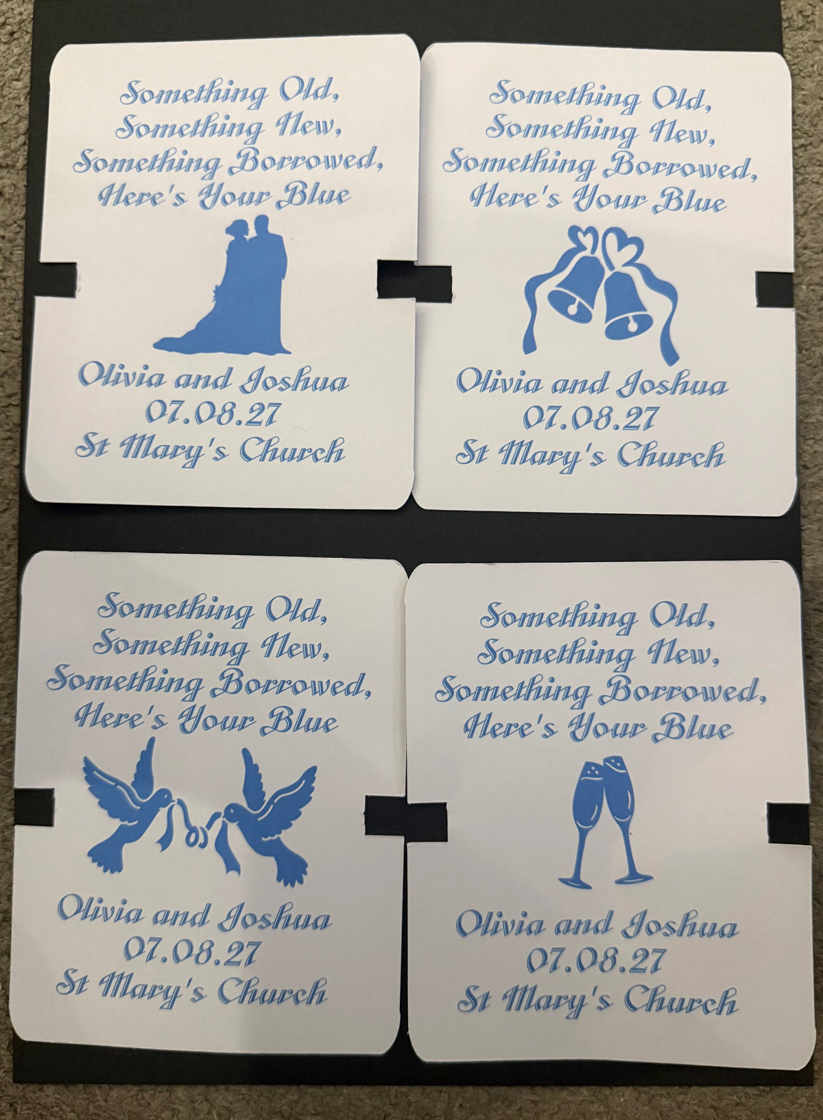 Four personalized wedding backing cards arranged in a 2x2 grid on a dark textured surface. Each coaster is white with blue printed text and a different wedding symbol: a bride and groom silhouette, bells with hearts, doves, and champagne glasses.