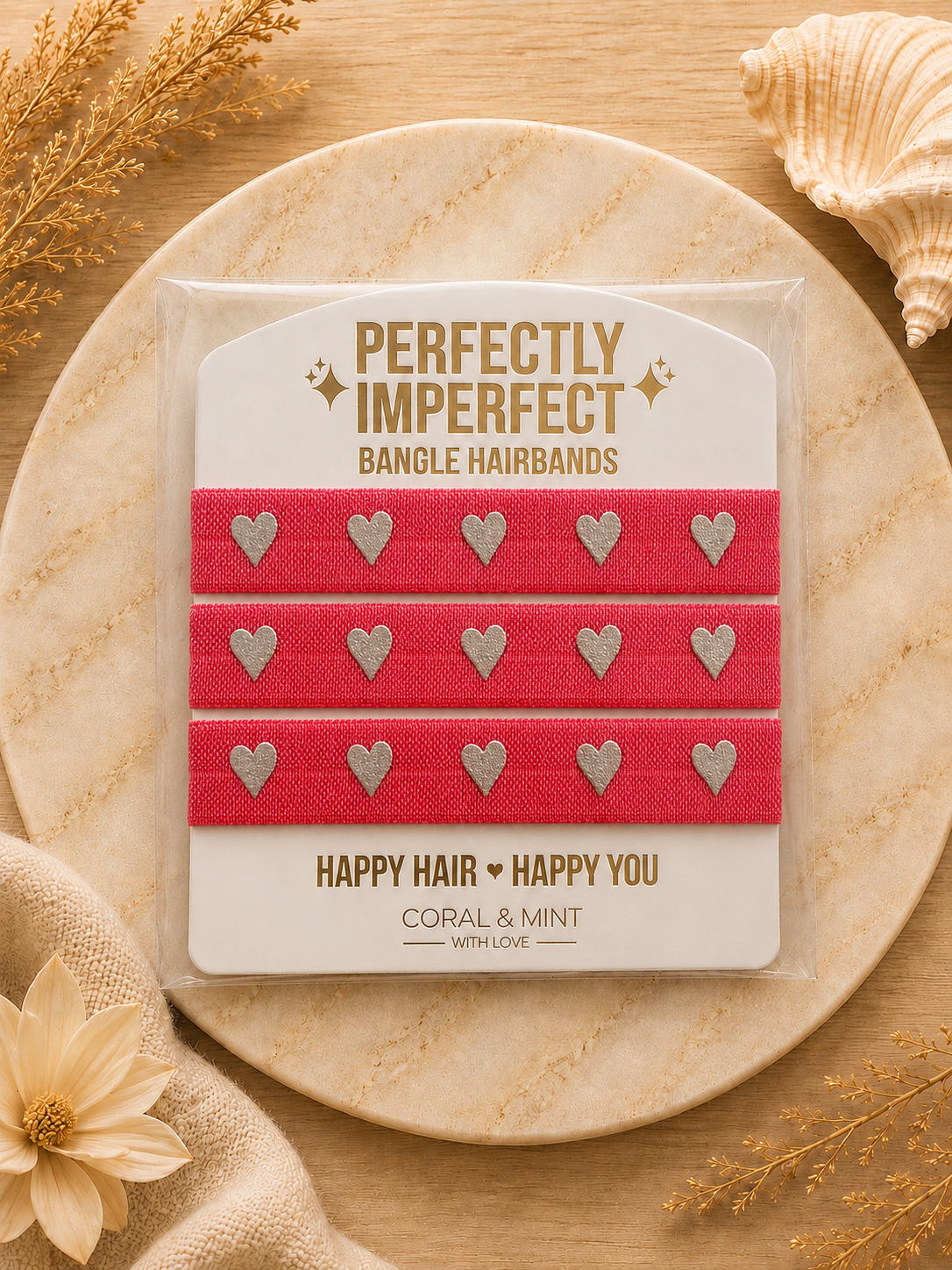 Three bright pink bangle hairbands with silver heart details, packaged on a branded card and displayed on a round wooden tray with decorative dried flowers and a seashell.