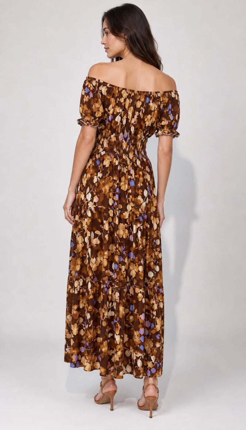A woman modeling an off-the-shoulder maxi dress with a brown, beige, and blue floral pattern, shown from the back against a light gray studio background.