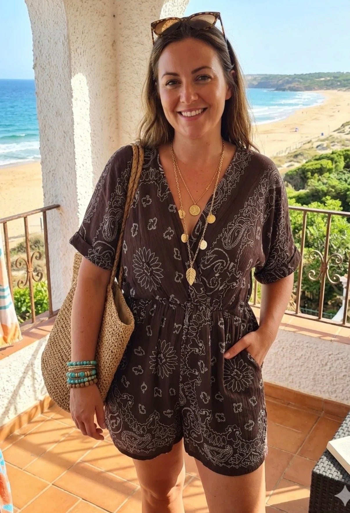 A woman stands on a sunlit balcony overlooking a beach, wearing a dark brown short-sleeve romper with a white paisley pattern, layered gold necklaces, sunglasses on her head, and a straw tote bag.