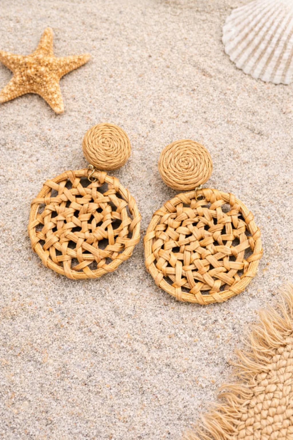Pair of brown woven earrings on a sandy surface with starfish and shells.