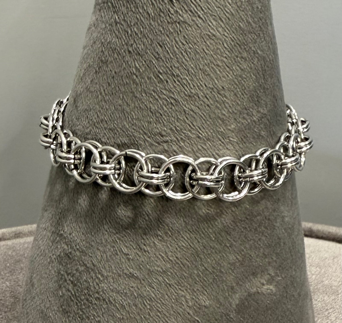 A silver chain link bracelet displayed on a gray textured cone stand against a neutral gray background.