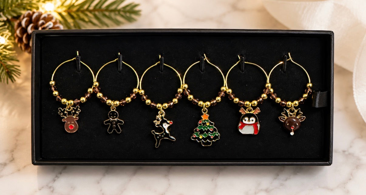 Six gold-tone wine glass charms with Christmas-themed pendants, including a reindeer, gingerbread man, Santa, Christmas tree, penguin, and reindeer head, displayed in a black presentation box on a marble surface with holiday greenery in the background.
