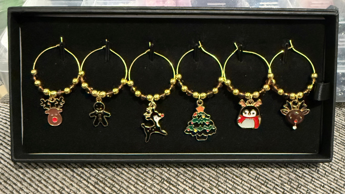 Six gold-tone wine glass charm hoops with Christmas-themed enamel pendants, displayed on a black presentation box.