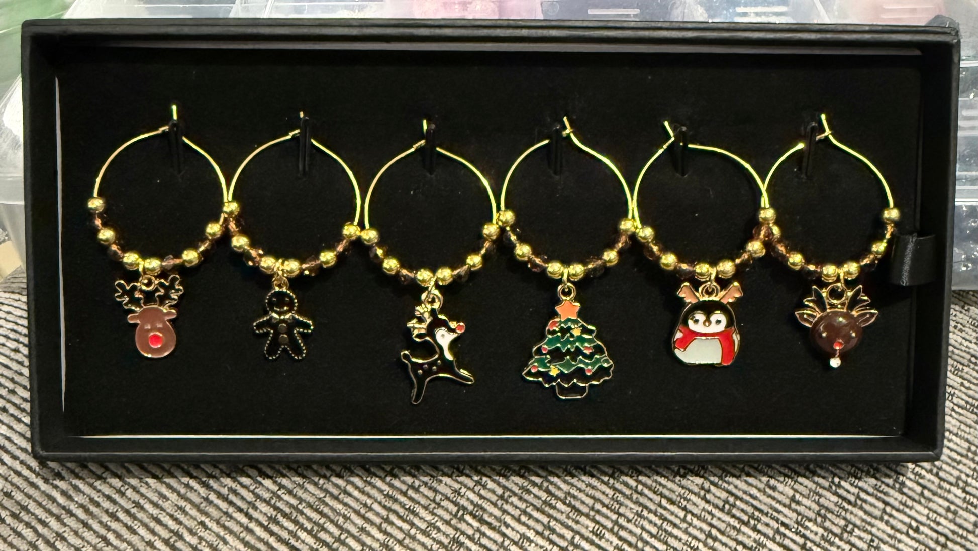 Six gold-tone wine glass charm hoops with Christmas-themed enamel pendants, displayed on a black presentation box.