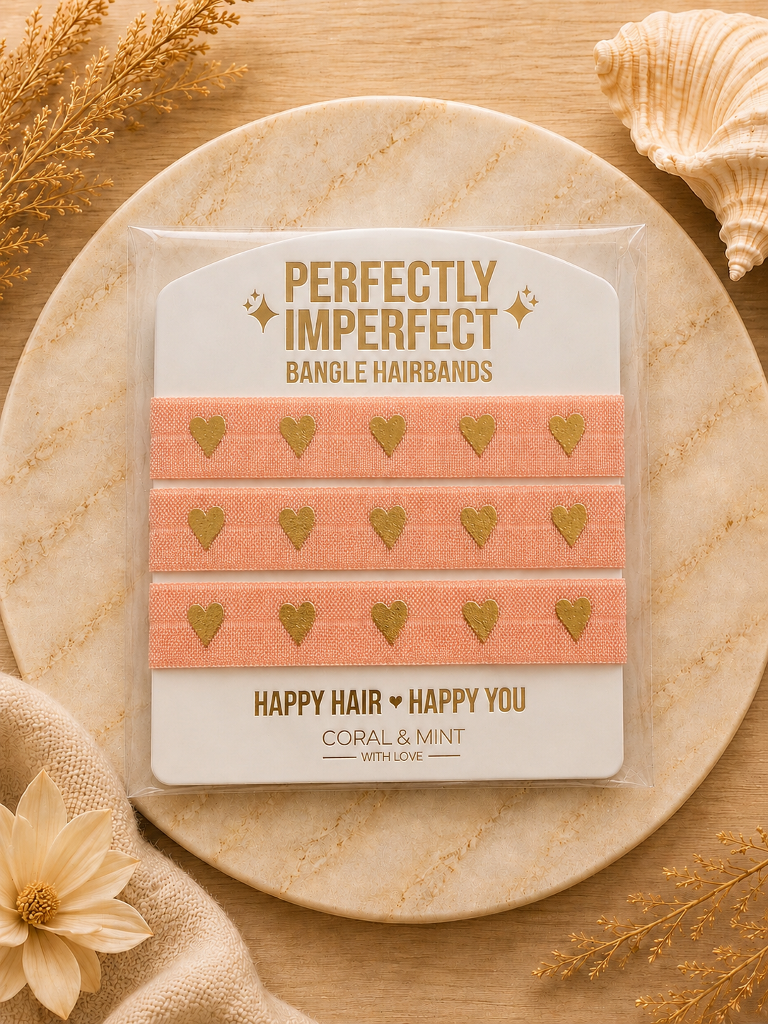 Three coral pink bangle hairbands with gold foil hearts, packaged on a branded card and displayed on a round stone tray with dried flowers and a seashell.