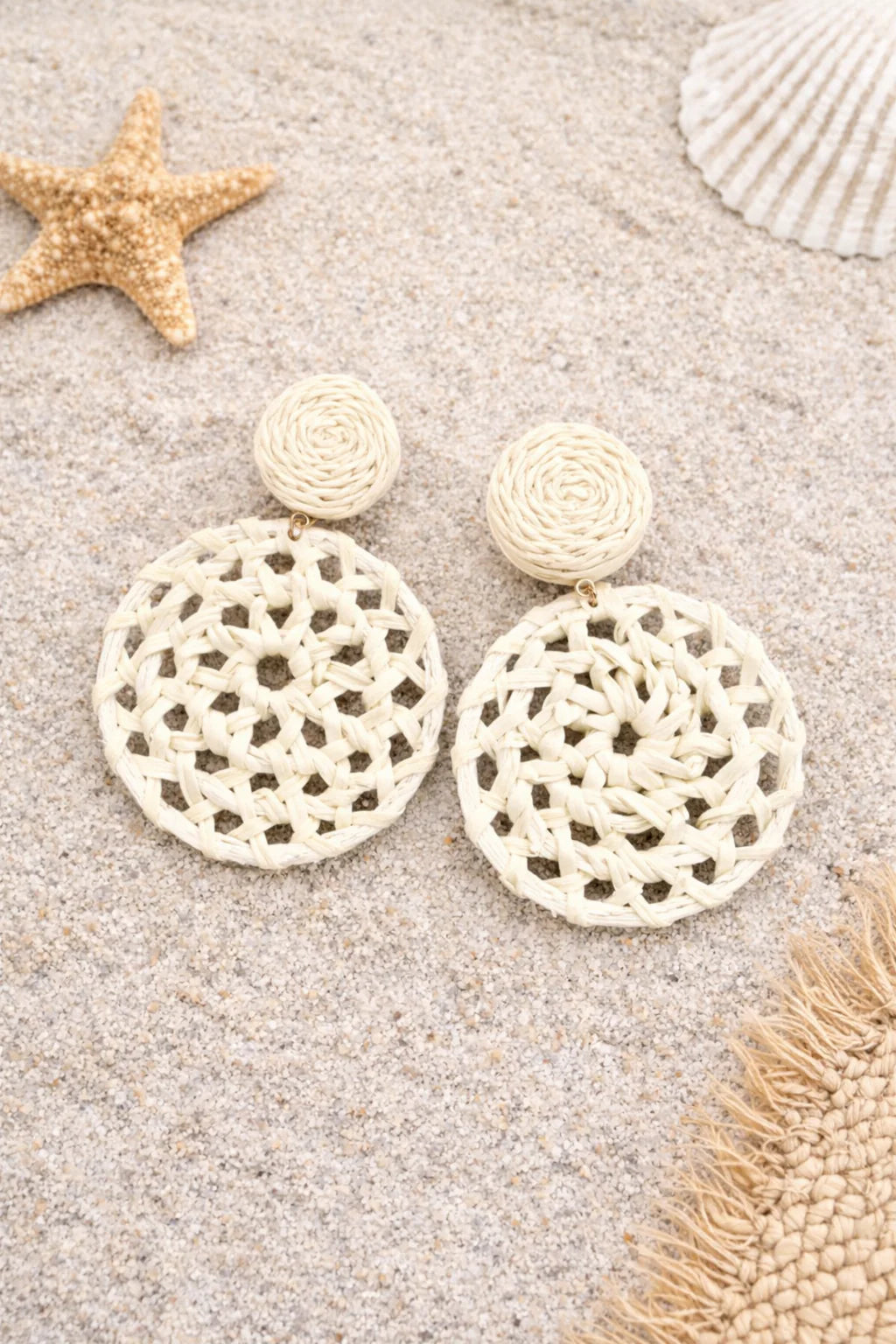Pair of white lace earrings on a sandy surface with starfish and shells.