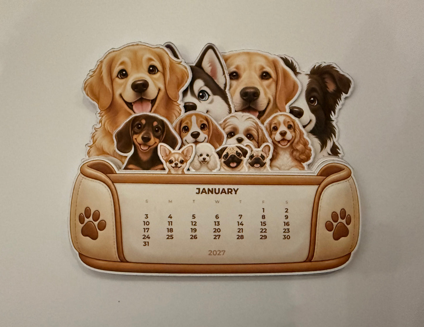 A novelty 2027 desk calendar featuring a collage of various dog breeds above a printed calendar grid. The calendar has a tan and brown design with paw print details on the sides.
