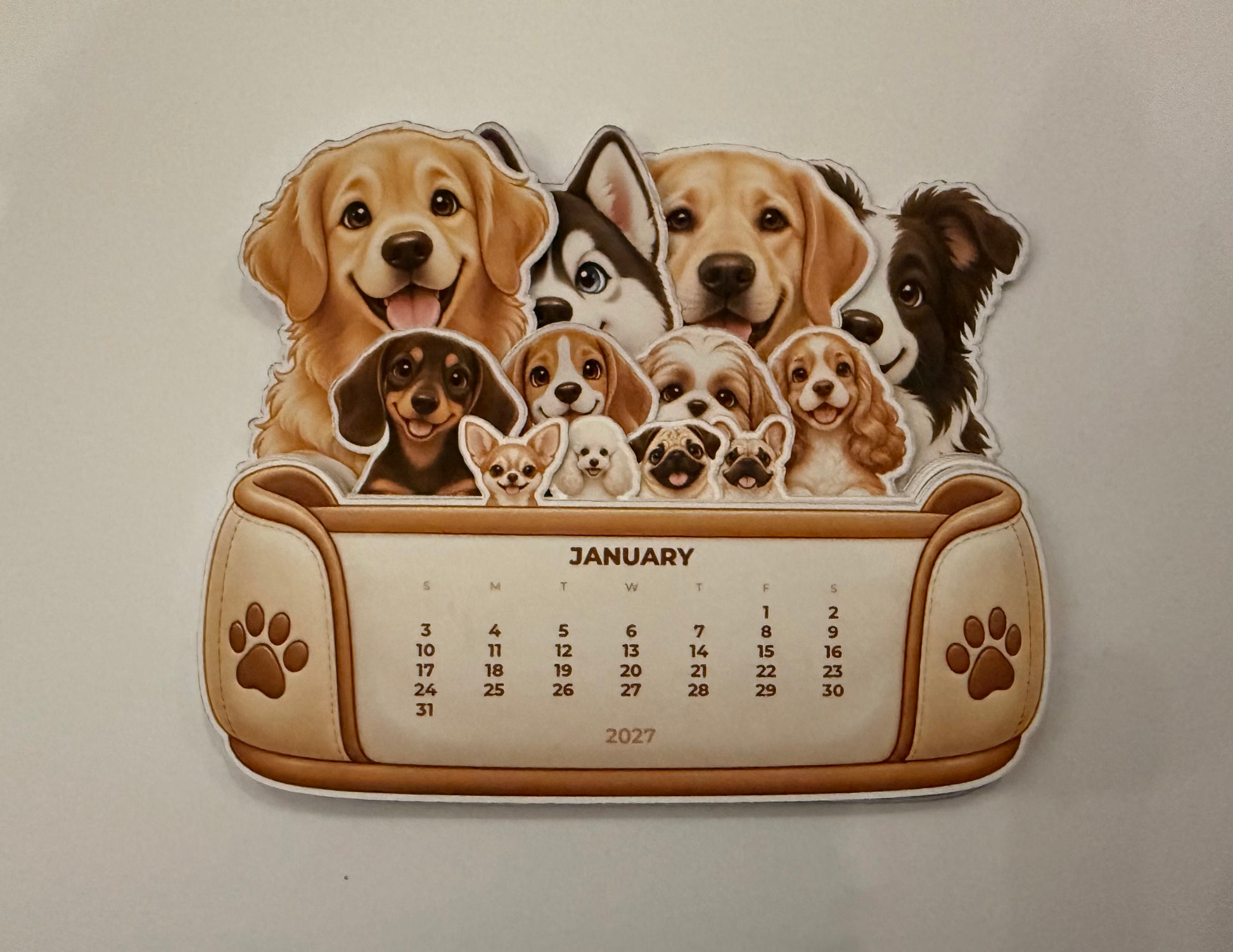 A novelty 2027 desk calendar featuring a collage of various dog breeds above a printed calendar grid. The calendar has a tan and brown design with paw print details on the sides.