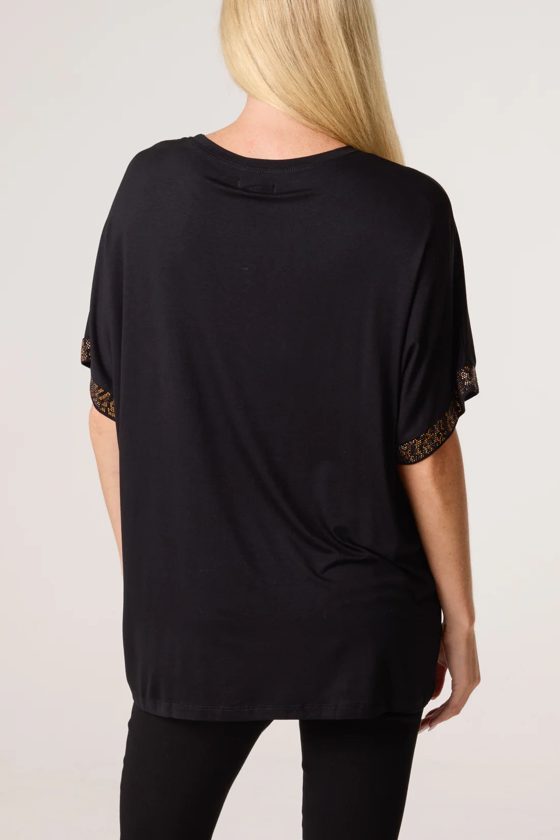 A woman viewed from the back wearing a loose black short-sleeve top with diamante trim at the sleeve cuffs, paired with black pants, against a light gray studio background.
