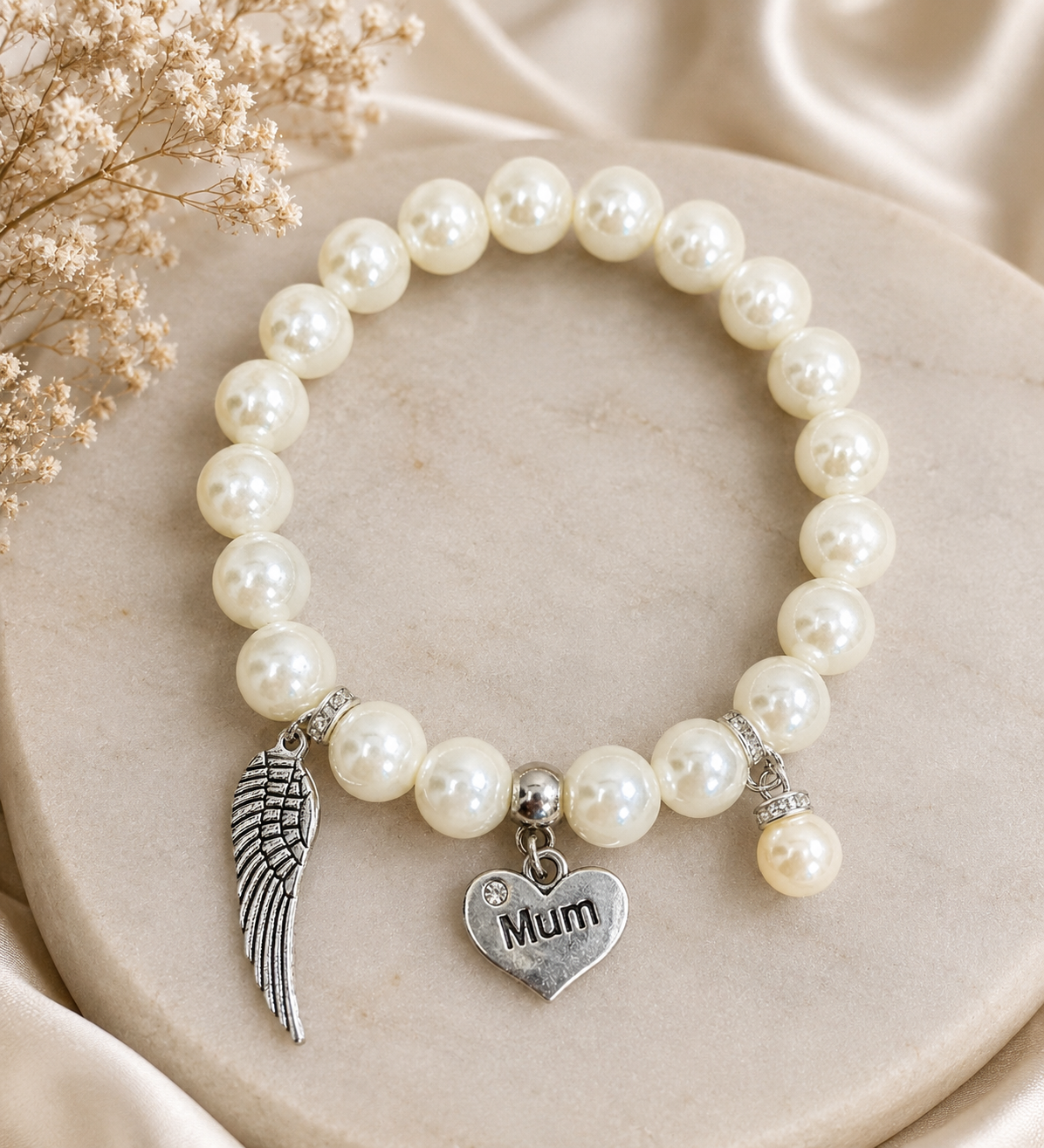 A pearl beaded bracelet with silver charms, including a wing charm and a heart charm engraved with 'Mum', displayed on a round stone slab with dried flowers and satin fabric.