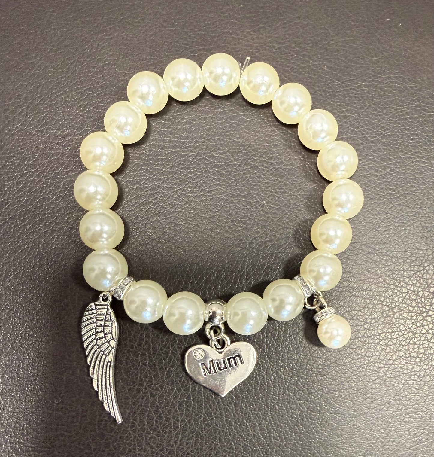 A beaded stretch bracelet with cream-colored faux pearls, featuring a silver angel wing charm, a silver heart charm engraved with 'Mum', and a small silver bead accent, displayed on a dark textured surface.