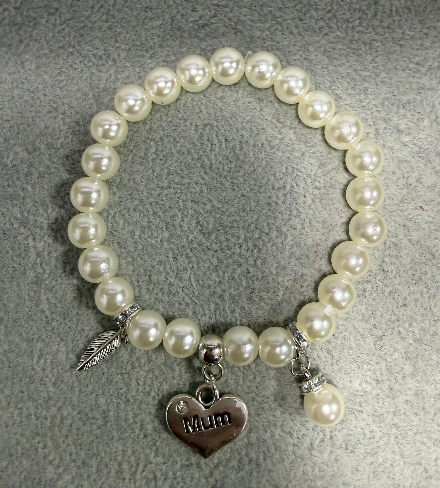 A pearl beaded stretch bracelet with silver-toned charms, including a heart-shaped pendant engraved with 'Mom' and a feather charm, displayed on a textured gray background.