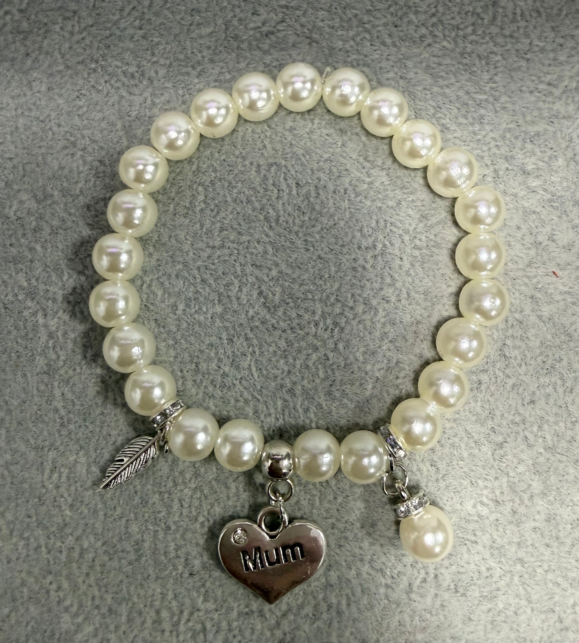 A pearl beaded stretch bracelet with silver-toned charms, including a heart-shaped pendant engraved with 'Mom' and a feather charm, displayed on a textured gray background.