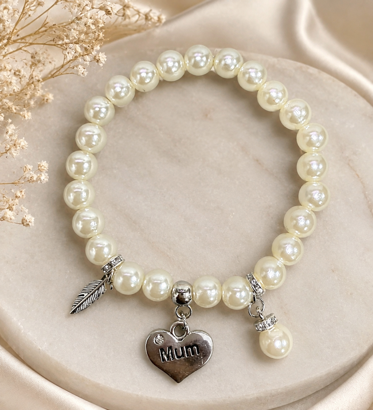 A beaded stretch bracelet featuring cream-colored faux pearls and silver-toned charms, including a heart-shaped charm engraved with 'Mum' and a feather, displayed on a beige marble surface with dried baby's breath flowers.