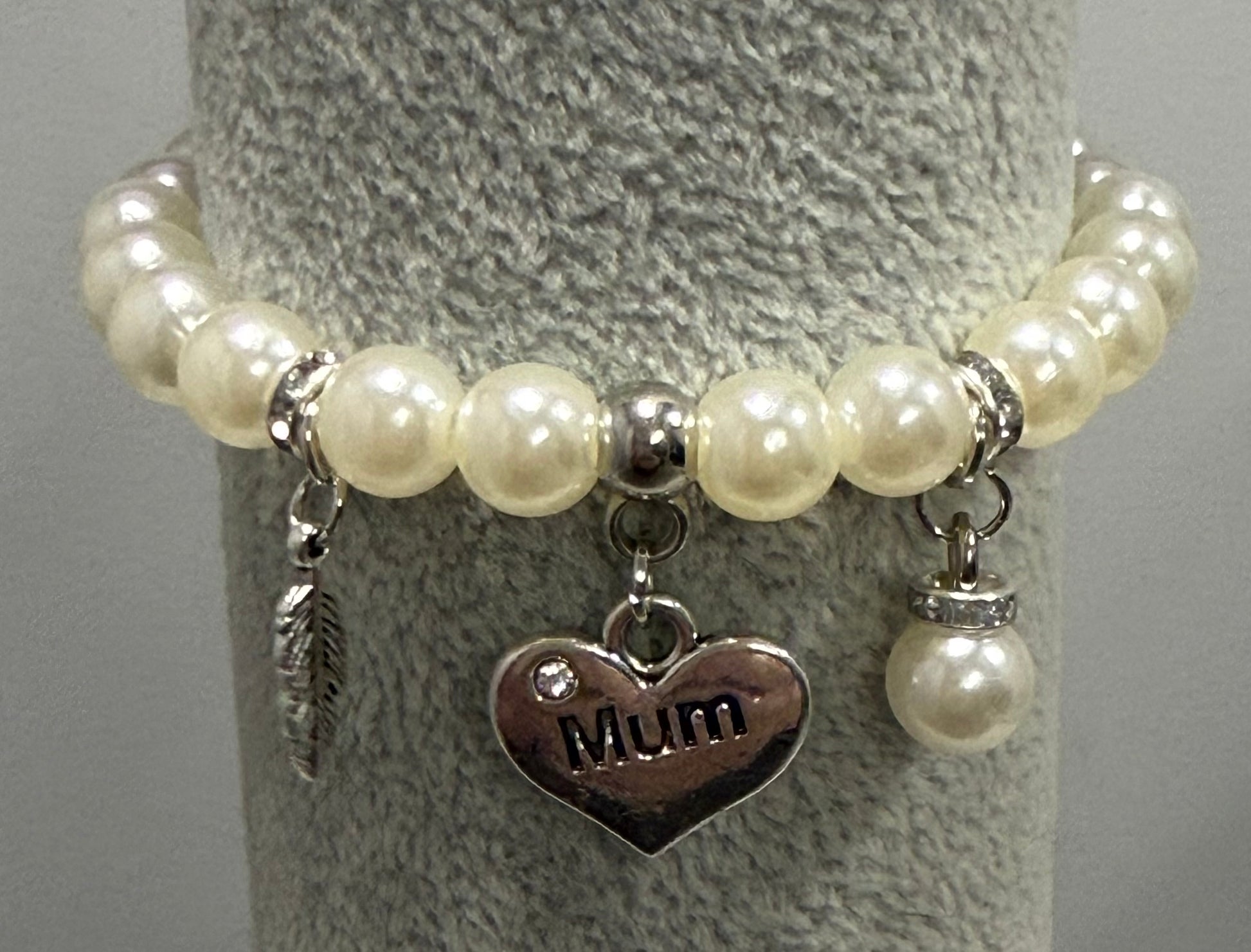 A beaded bracelet with cream-colored faux pearls, silver-toned spacer beads, and three charms: a feather, a heart engraved with 'Mum', and a small pearl with a rhinestone cap, displayed on a gray textured stand.