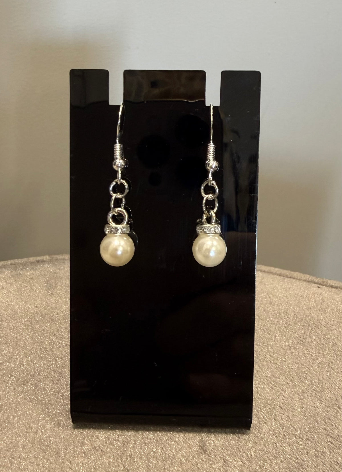 A pair of silver-toned dangle earrings with white pearl drops, displayed on a black stand against a neutral background.