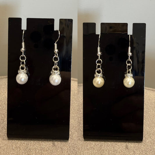 A pair of dangling earrings displayed on black stand cards. Each earring has a silver-tone fishhook and chain with a small clear rhinestone cap and a white pearl drop.