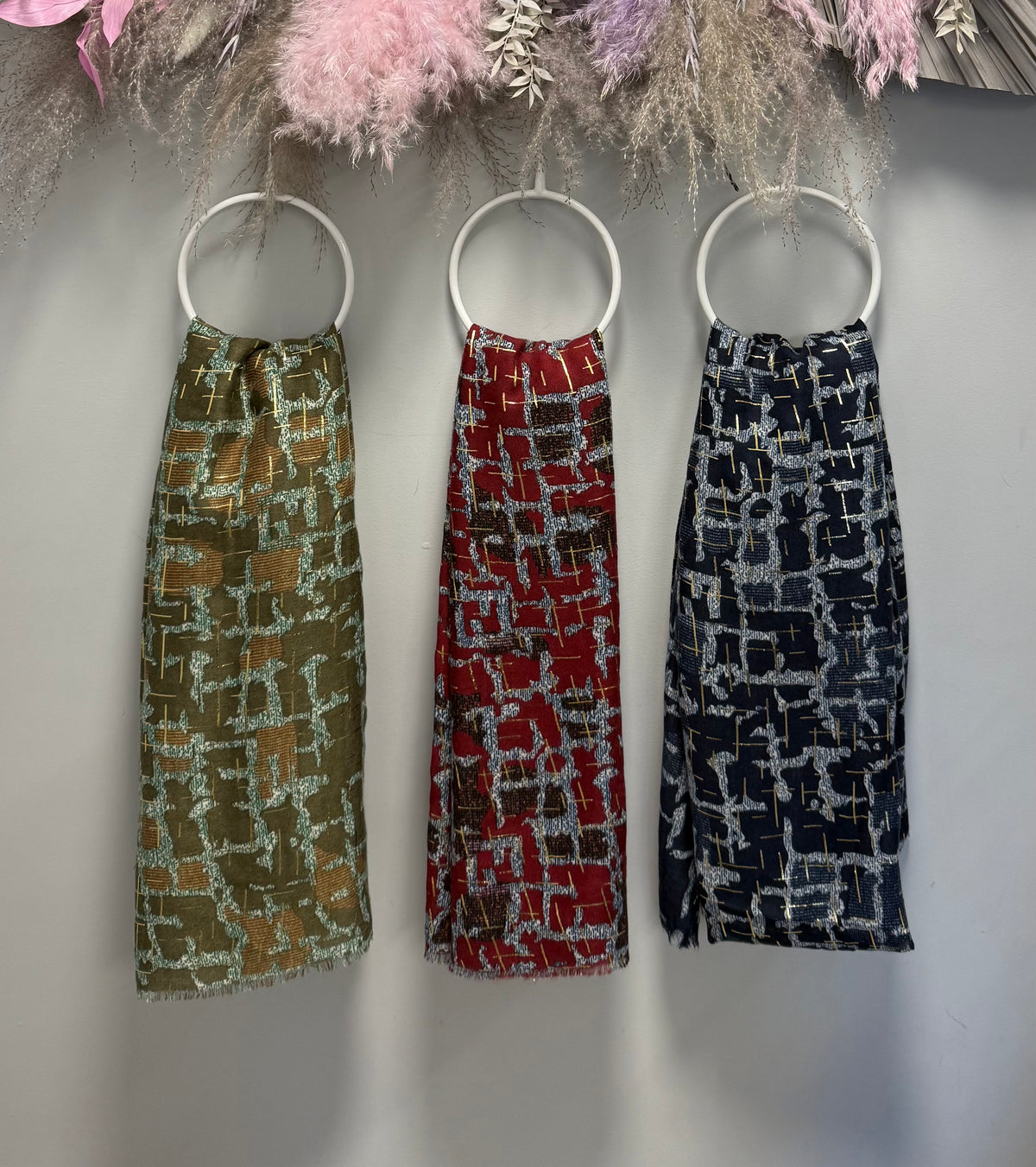 Three gold foil detail scarfs in olive green, red, and navy blue hang on white circular rings against a light gray wall with dried floral decorations at the top.