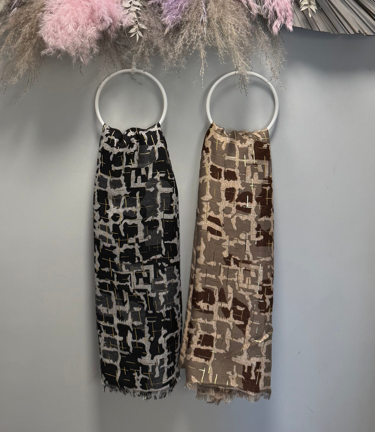 Two patterned gold foil detail scratchs hanging on white circular rings against a gray wall, with dried floral decor at the top. One dress is black and white, and the other is brown and beige.