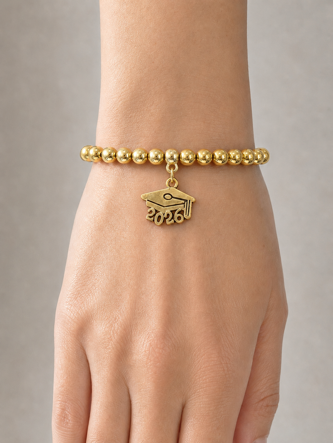 A gold beaded bracelet worn on a person's wrist, featuring a small graduation cap charm with the year 2026 engraved on it, shown against a neutral gray background.