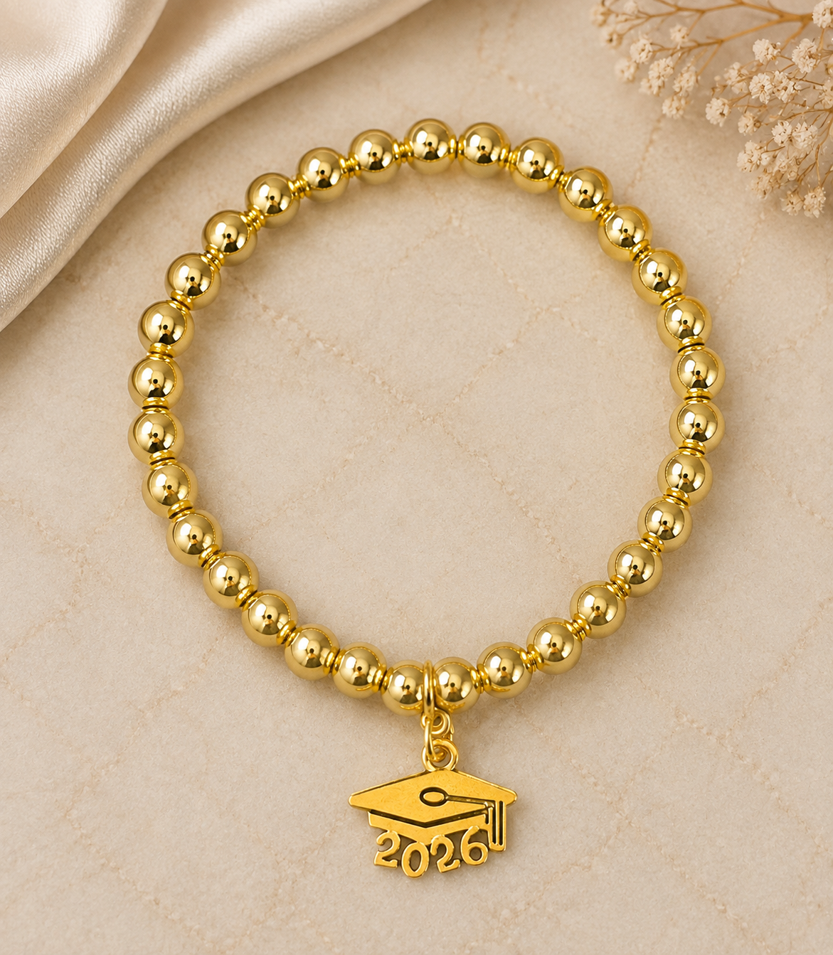 A gold beaded bracelet with a graduation cap charm engraved with '2026', displayed on a beige textured surface with a satin fabric and dried flowers in the background.