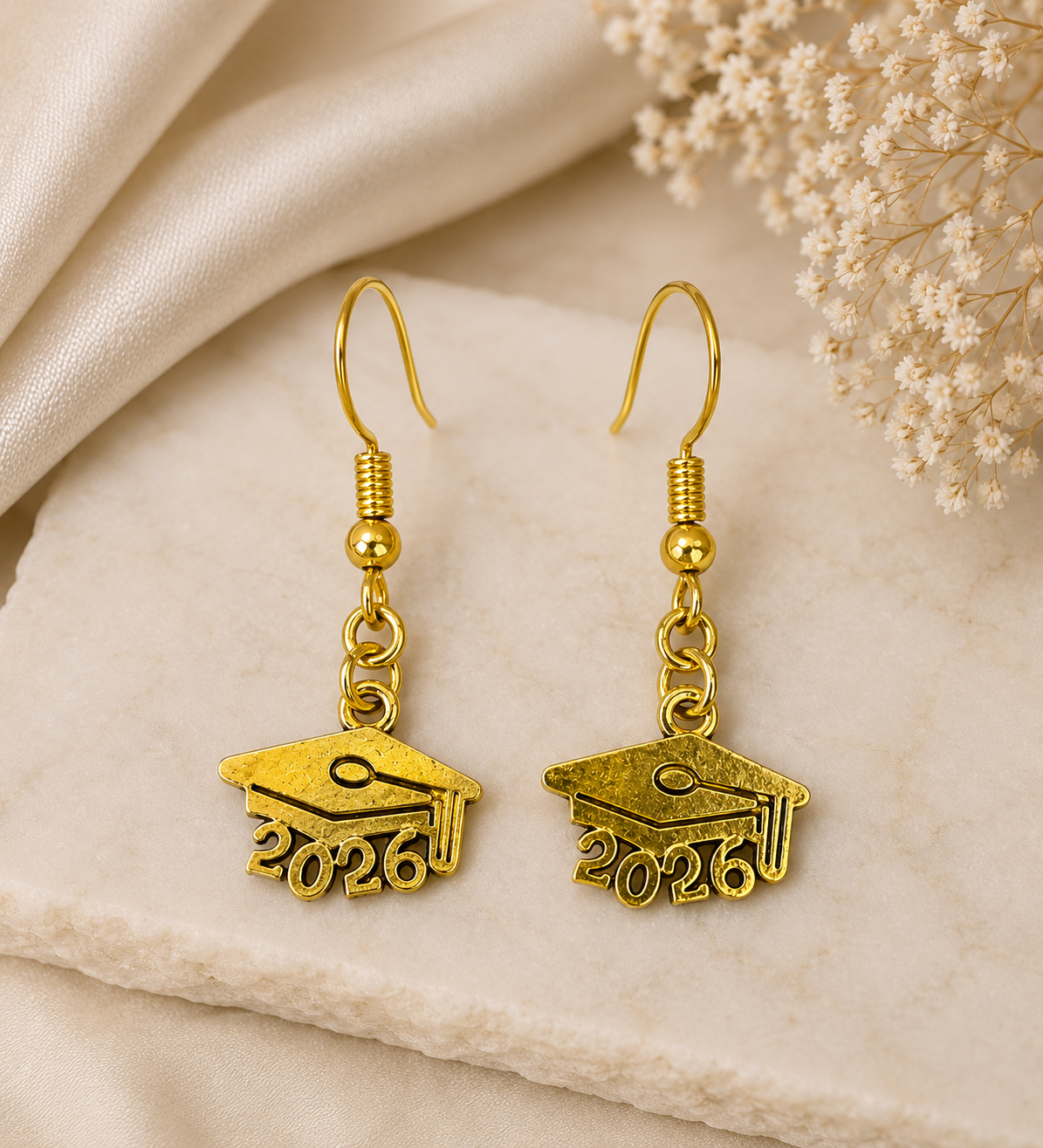 A pair of gold drop earrings shaped like graduation caps with the year '2026' engraved on the front, displayed on a textured cream stone with draped satin fabric and dried flowers in the background.