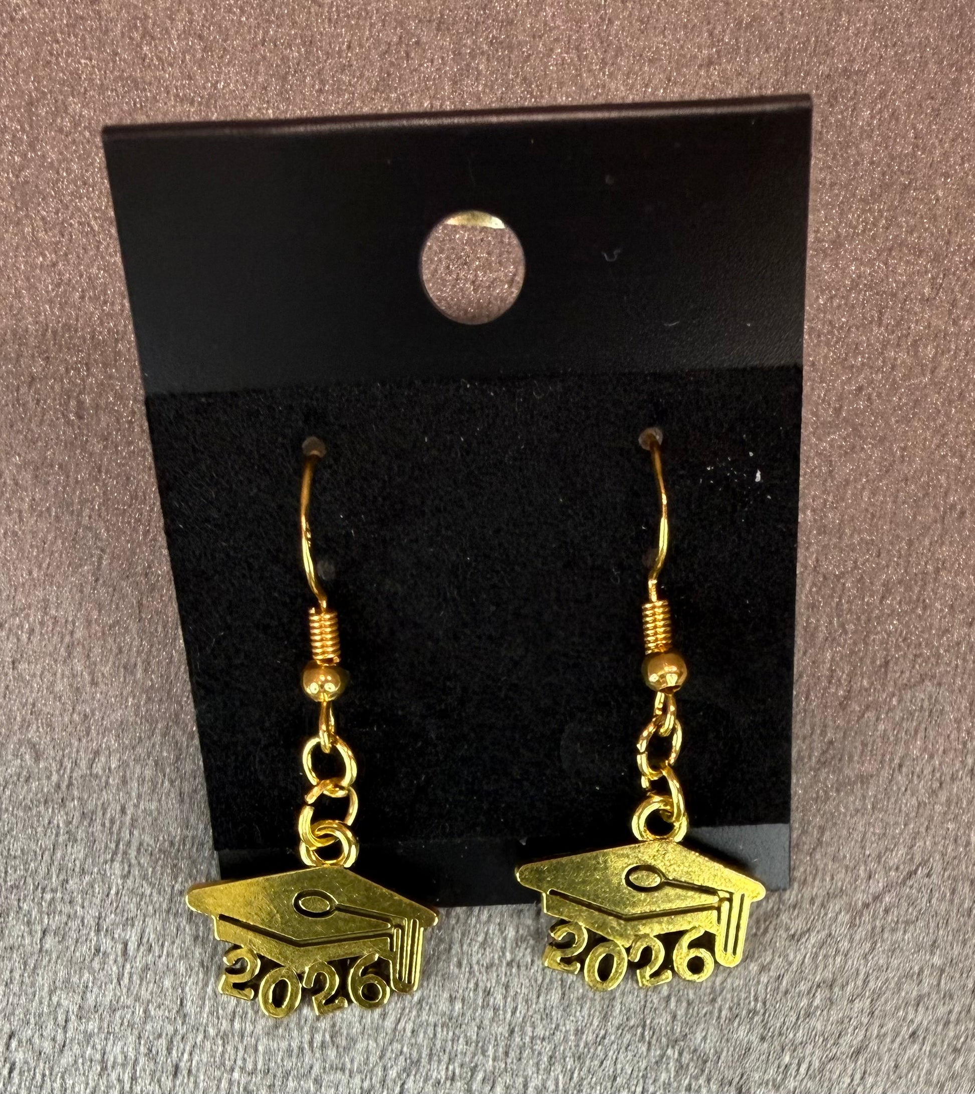 A pair of gold-colored dangle earrings shaped like graduation caps with the year '2025' engraved on the bottom, displayed on a black velvet card against a textured gray background.