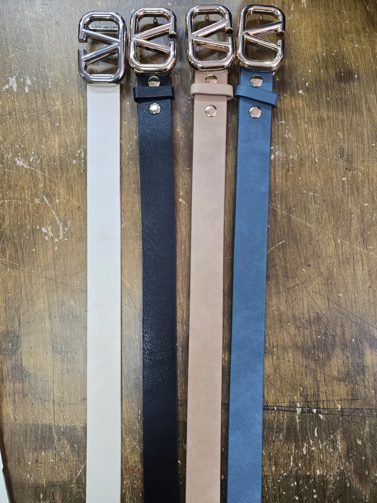 Four belts in white, black, beige, and blue with gold buckles on a wooden surface.