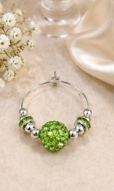 A silver wine glass charm with a large green faceted bead and smaller green and silver beads, displayed on a beige fabric surface with small white flowers and a soft satin cloth in the background.