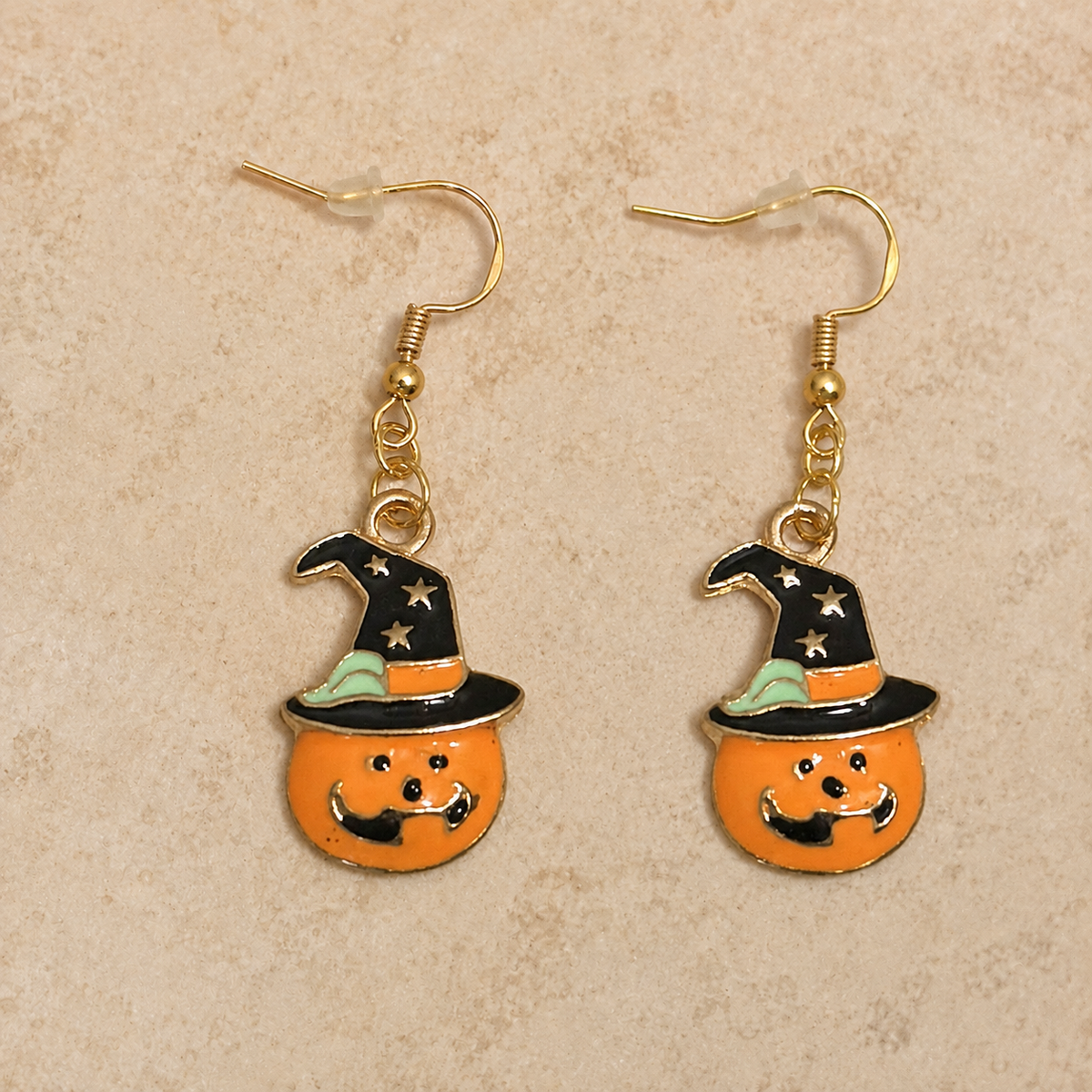 A pair of gold-tone dangle earrings featuring orange pumpkin charms with black witch hats, displayed on a textured beige background.