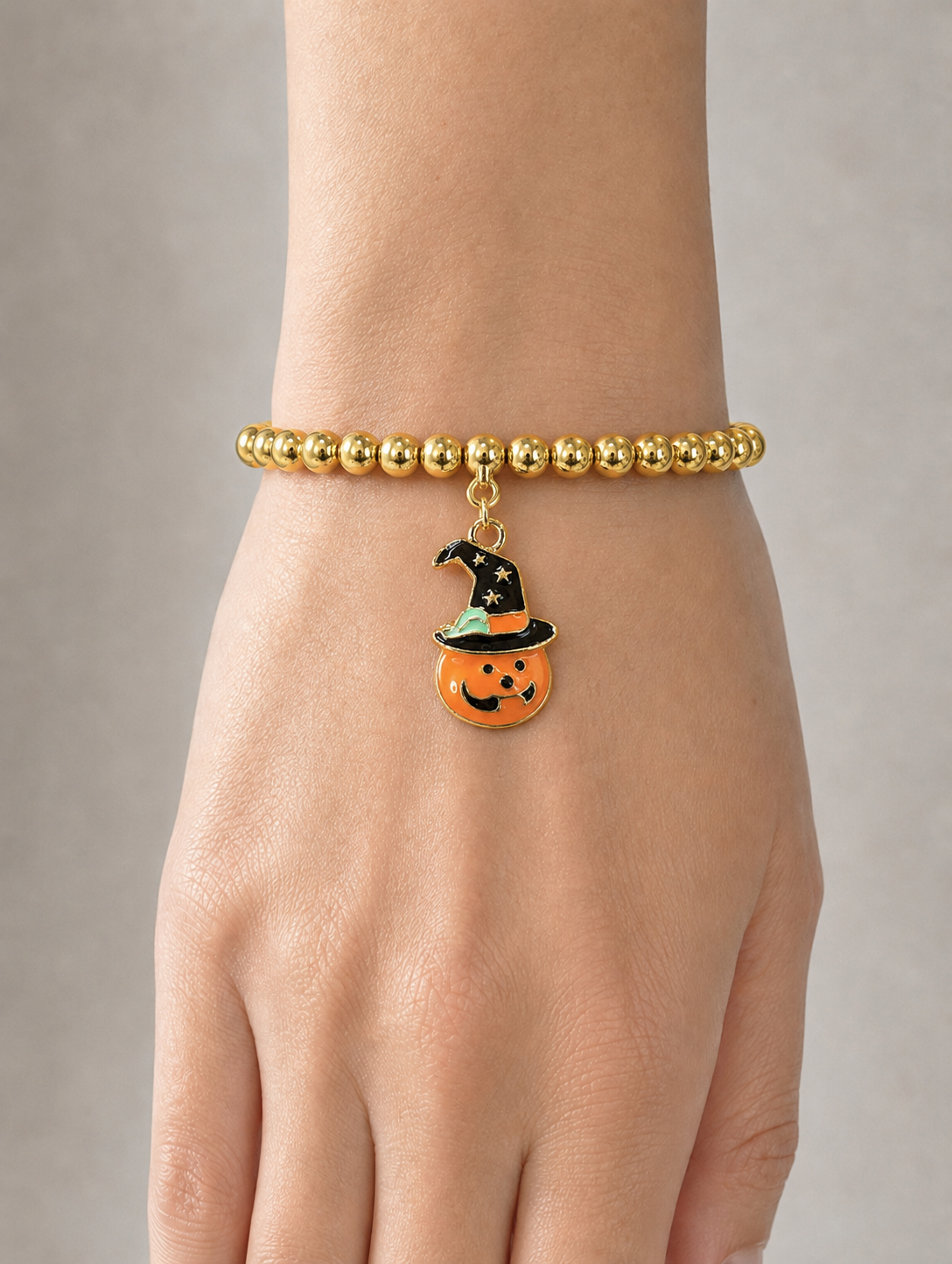 A gold beaded bracelet worn on a wrist, featuring a dangling orange pumpkin charm with a black witch hat.