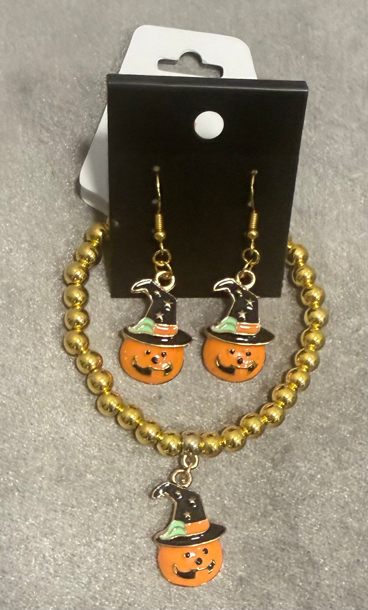 A pair of gold beaded drop earrings with orange pumpkin charms wearing black witch hats, displayed on a black card hanger against a gray textured background.