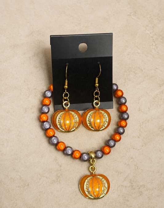 A pair of drop earrings and a matching beaded necklace displayed on a black card hanger against a beige textured background. The jewelry features orange pumpkin-shaped charms with gold trim, connected to strands of orange, gray, and gold-toned beads.