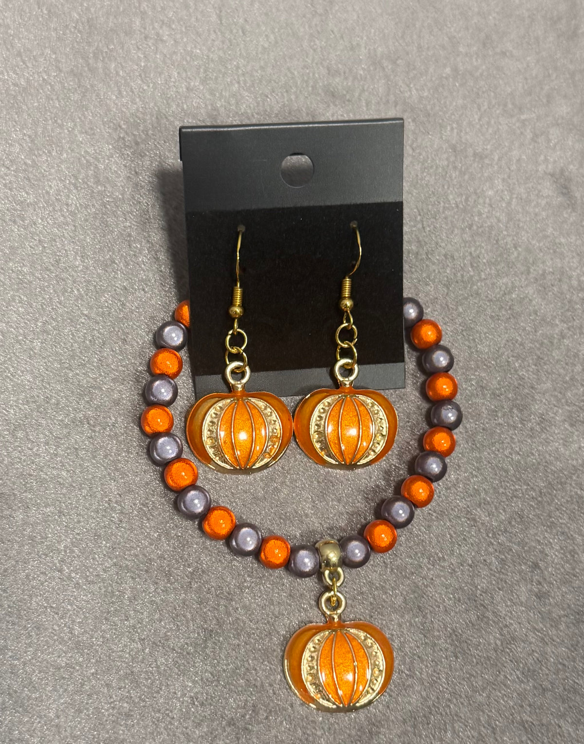 A pair of beaded drop earrings displayed on a black card against a gray textured background. The earrings feature orange and gray beads with gold-tone hooks and oval orange-and-gold pumpkin charms.