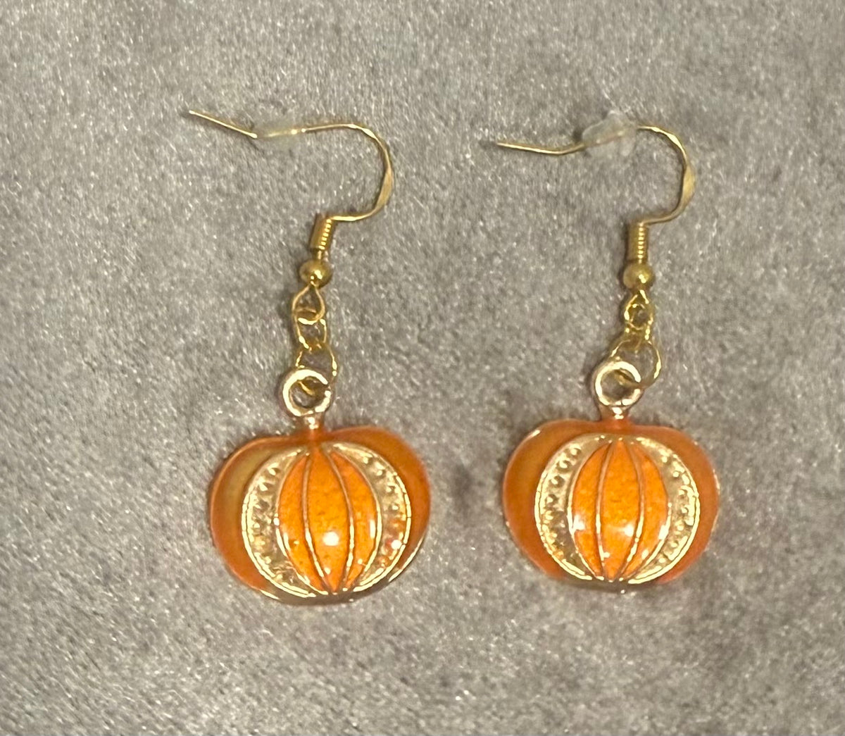 A pair of gold-tone dangle earrings with orange pumpkin-shaped charms, displayed on a textured gray background.