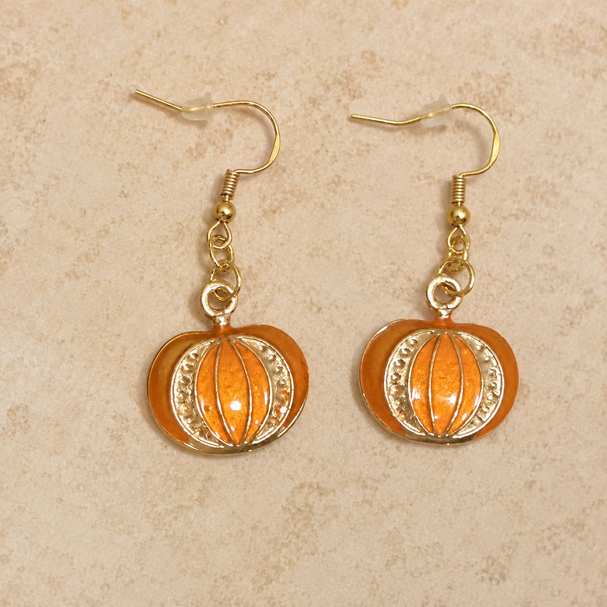 A pair of gold-tone dangle earrings with orange pumpkin charms, displayed on a textured beige background.